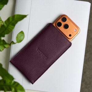 May include: A deep burgundy leather phone sleeve with a textured surface. A gold-colored smartphone is partially visible inside the sleeve. The sleeve is resting on a white surface, with green foliage in the background.