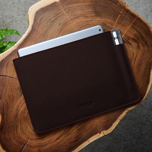 Könnte beinhalten: Eine dunkelbraune Tablet-Hülle aus Leder mit einem teilweise eingesetzten silbernen Tablet. Ein weißer Stift befindet sich in einer Filztasche. Die Hülle liegt auf einer Holzoberfläche mit natürlicher Kante. Die Gesamtästhetik ist minimalistisch und modern.