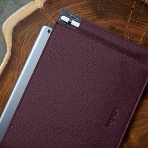 May include: A burgundy leather tablet sleeve with a silver tablet partially inserted. A white stylus with black accents is in a gray fabric pocket. The sleeve is on a dark wood surface. The word "voyage" is embossed on the leather.