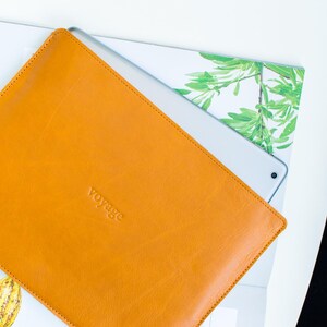 Könnte beinhalten: Eine orangefarbene Tablet-Hülle aus Leder mit dem Wort "voyage" auf der Vorderseite. Ein silbernes Tablet ist teilweise sichtbar. Der Hintergrund zeigt eine weiße Oberfläche und einen Aufdruck mit grünen Blättern. Die Hülle ist geöffnet.