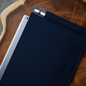Puede incluir: Una funda de tableta azul marino con una superficie texturizada, que contiene una tableta plateada. Un lápiz óptico blanco está en un bolsillo de fieltro gris. La funda está sobre una superficie de madera.