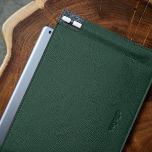 May include: A dark green leather tablet sleeve with a silver tablet partially inserted. A white and black stylus is in a grey felt pocket at the top. The sleeve is on a dark wood surface.