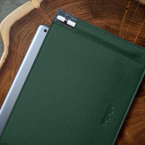 May include: A dark green leather tablet sleeve with a silver tablet partially inserted. A white and gray stylus is in a holder at the top. The sleeve has the word "voyage" embossed on the lower right corner. The sleeve is on a wooden surface.