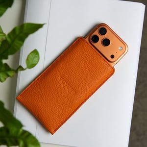 以下が含まれることがあります： オレンジ色の革製携帯電話ケースに、金色の携帯電話が部分的に挿入されています。ケースは質感のある表面で、前面にはブランド名「Vovoo」がエンボス加工されています。携帯電話には3つのカメラレンズがあります。