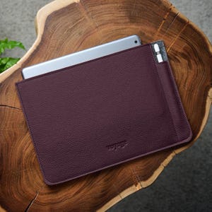 Pode incluir: Uma capa de tablet de couro bordô com uma superfície texturizada. A capa tem um bolso para uma caneta stylus e a palavra "voyage" em relevo na frente. Um tablet prateado é parcialmente visível.