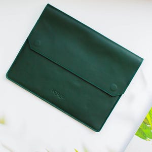 May include: A dark green leather laptop sleeve with a button closure. The word "voyage" is embossed on the lower front. The sleeve is rectangular and has a flap that folds over to secure the contents.