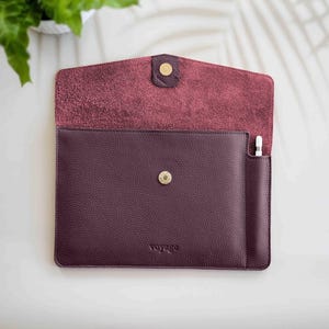 May include: A burgundy leather tablet sleeve with a flap closure and a gold snap. The interior is a lighter shade of burgundy. A small pocket holds a white stylus. The word "voyage" is embossed on the front.