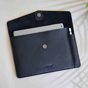 Könnte beinhalten: Eine schwarze Lederhülle für iPad mit Klappverschluss und Druckknopf. Die Hülle hat ein Fach für einen Stift und ein Logo auf der Vorderseite.