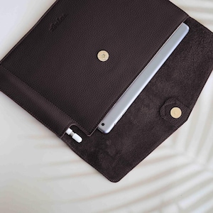 Puede incluir: Una funda de tableta de cuero marrón oscuro con un cierre de botón a presión dorado. La funda está abierta, revelando una tableta plateada y un lápiz óptico blanco. La funda tiene una superficie texturizada y un interior suave.