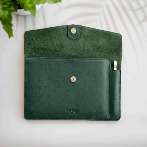 Könnte beinhalten: Dunkelgrüne Tablet-Hülle aus Leder mit Klappenverschluss und goldenem Druckknopf. Die Hülle hat eine Tasche für einen Stift und das Wort "VOYAGE" ist unten eingeprägt. Die Innenseite ist in einem helleren Grünton gehalten.