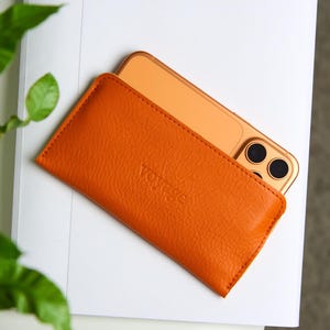 Puede incluir: Funda de teléfono de cuero naranja con la palabra "voyage" grabada en relieve en la parte delantera. Un teléfono inteligente de color dorado es parcialmente visible dentro de la funda. La funda es rectangular y tiene una superficie texturizada. La funda del teléfono está sobre una superficie blanca.
