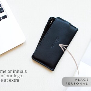 May include: Black leather phone case with the word "voyage" embossed on the front. The case is on a white surface next to a laptop. Text on the image says "Your name or initials instead of our logo. Available at extra charge!" and "PLACE FOR PERSONALIZATION."