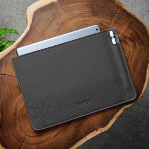 Könnte beinhalten: Eine dunkelgraue Tablet-Hülle aus Leder mit einem teilweise eingelegten silbernen Tablet. Die Hülle hat eine strukturierte Oberfläche und eine kleine Tasche für einen Stift. Das Wort "voyage" ist in der unteren rechten Ecke geprägt.