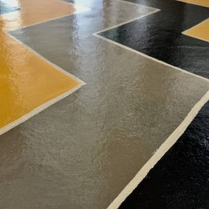 Può includere: Primo piano di un pavimento con un motivo a chevron dipinto in nero, grigio e giallo. Il pavimento è lucido e riflette la luce.