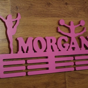 Può includere: Espositore per medaglie in legno rosa con il nome "MORGAN" e due figure di cheerleader. L'espositore ha cinque slot per le medaglie.