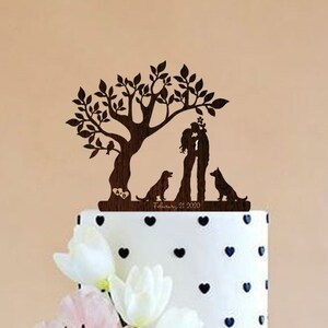 May include: A three-tiered wedding cake decorated with black hearts on a white background. The cake is topped with a wooden cake topper featuring a couple kissing under a tree with two dogs. The date "February 21, 2020" is engraved on the cake topper.