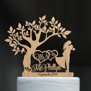 May include: A white wedding cake with a pink ribbon and white flowers on top. The cake is decorated with a wooden topper featuring a tree with two figures standing under it. The topper reads "Mr & Mrs Phillips 1 September 2017".