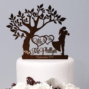 May include: A two-tier wedding cake decorated with white flowers, pine cones, and berries. The cake topper is a wooden silhouette of a couple standing under a tree with the text "Mr. & Mrs. McPhillips 1 September 2017".