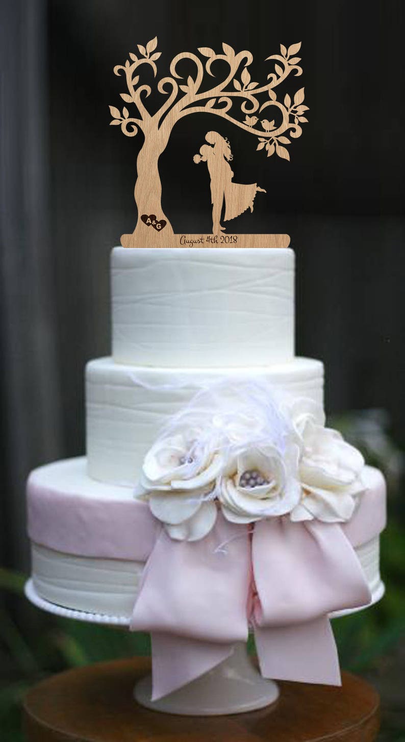 Puede incluir: Un adorno de pastel de boda de madera con una pareja bes&aacute;ndose bajo un &aacute;rbol con p&aacute;jaros. El adorno est&aacute; sobre un pastel blanco con glaseado rosa y un lazo blanco. El texto "August 4th 2018" est&aacute; grabado en el adorno.