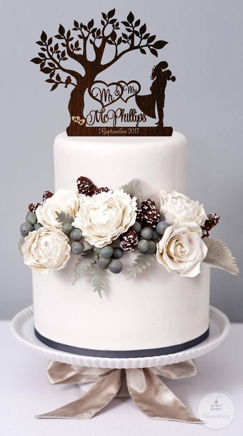 May include: A two-tier wedding cake decorated with white flowers, pine cones, and berries. The cake topper is a wooden silhouette of a couple standing under a tree with the text "Mr. & Mrs. McPhillips 1 September 2017".