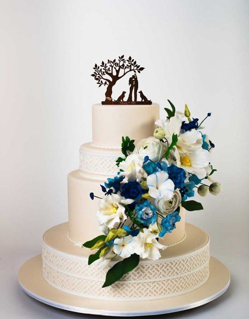 May include: A three-tiered wedding cake decorated with white fondant and a delicate lace pattern. The cake is adorned with a cascading arrangement of white, blue, and green sugar flowers. A wooden topper features a silhouette of a couple kissing under a tree with two dogs.