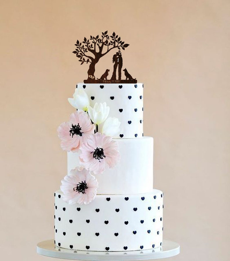 May include: A three-tiered wedding cake decorated with black hearts on a white background. The cake is topped with a wooden cake topper featuring a couple kissing under a tree with two dogs. The date "February 21, 2020" is engraved on the cake topper.