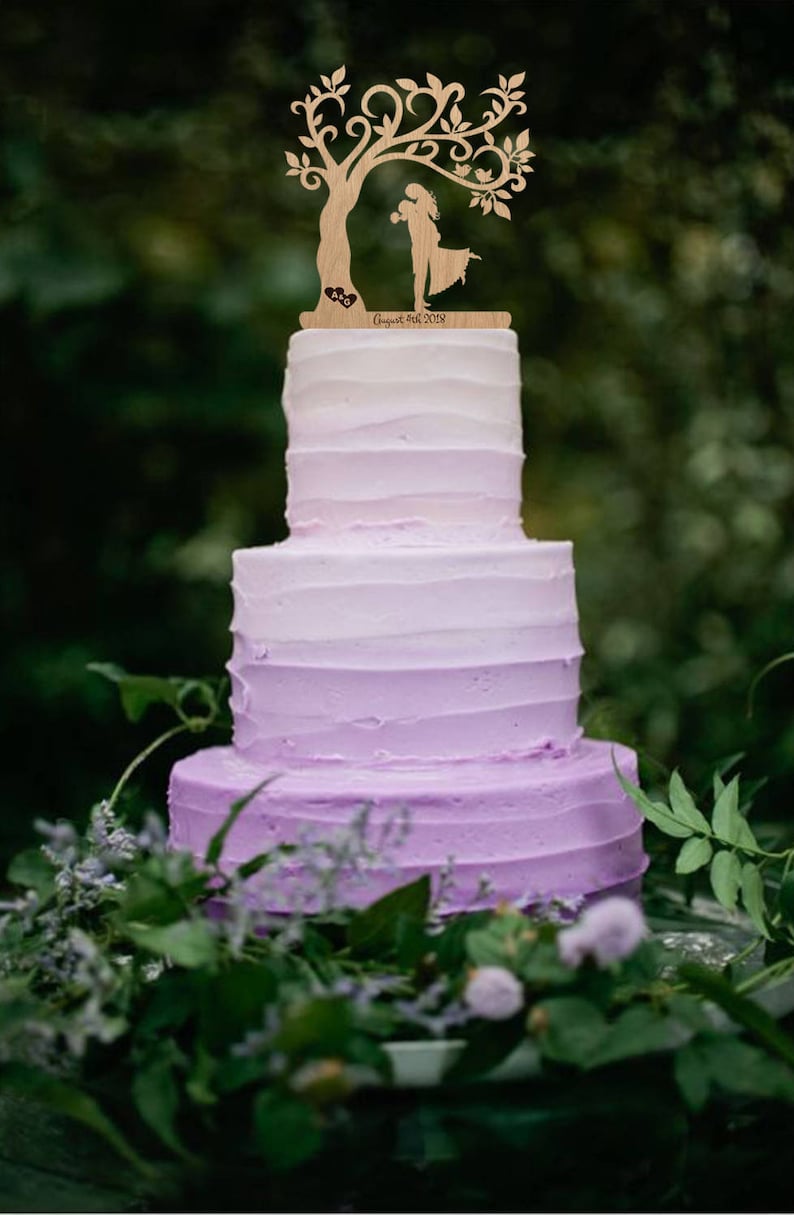 Puede incluir: Un pastel de boda de tres niveles con glaseado morado degradado. El pastel est&aacute; decorado con un adorno de pastel de madera que presenta una pareja bes&aacute;ndose debajo de un &aacute;rbol con un p&aacute;jaro posado en una rama. El adorno tambi&eacute;n incluye el texto "August 18th, 2018".