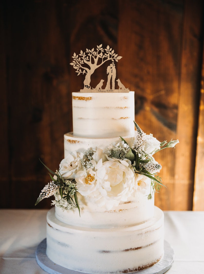 May include: A three-tiered white wedding cake with a wooden topper featuring a couple and two dogs under a tree. The cake is decorated with white flowers and greenery.