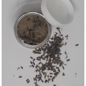 May include: A small glass jar with a silver lid filled with a brown, lavender-infused cream. Dried lavender flowers are scattered around the jar on a white surface.