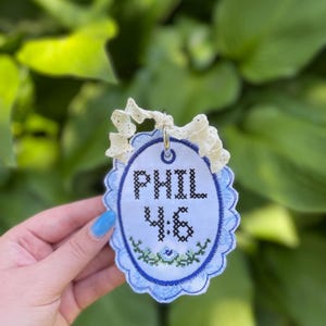 K&ouml;nnte beinhalten: Ein ovales Stoffornament mit dem in Kreuzstich gestickten schwarzen Text PHIL 4:6 &uuml;ber einem kleinen gr&uuml;n-blauen Blumenmotiv. Das Ornament hat einen gewellten blauen Rand und ist an einer cremefarbenen Spitzenbandschlaufe befestigt.