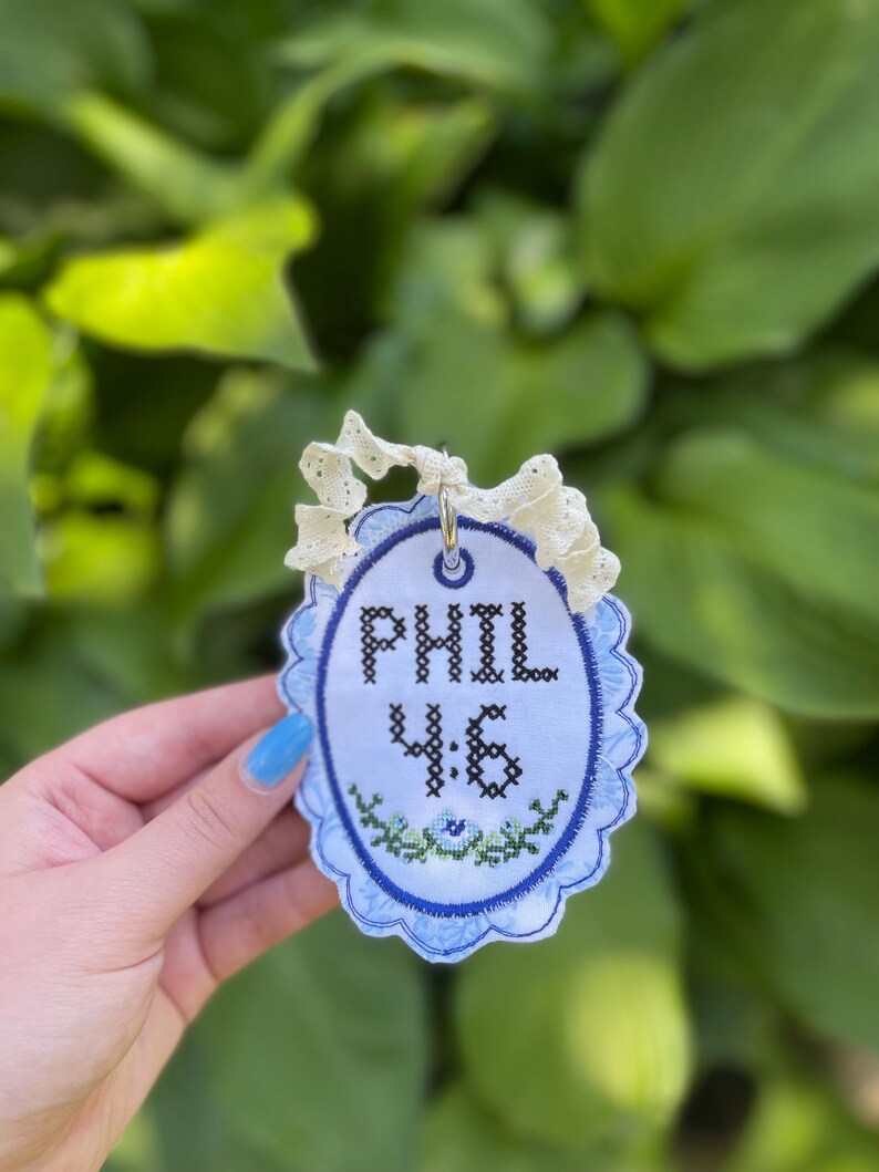 K&ouml;nnte beinhalten: Ein ovales Stoffornament mit dem in Kreuzstich gestickten schwarzen Text PHIL 4:6 &uuml;ber einem kleinen gr&uuml;n-blauen Blumenmotiv. Das Ornament hat einen gewellten blauen Rand und ist an einer cremefarbenen Spitzenbandschlaufe befestigt.