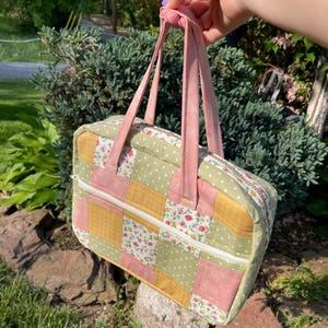 Puede incluir: Una bolsa de mano de patchwork con tela rosa, verde y amarilla. La bolsa tiene cierre de cremallera y asas de tela rosa.