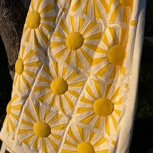 Peut inclure: Une courtepointe blanche avec un motif de soleil jaune. La courtepointe est faite de carrés avec un soleil jaune au centre de chaque carré. Les rayons du soleil sont matelassés avec du fil blanc.