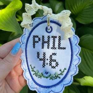 May include: An oval-shaped fabric ornament featuring the black cross-stitched text PHIL 4:6 above a small green and blue floral embroidery. The white fabric has a scalloped blue border and is topped with a cream-colored eyelet lace ribbon.