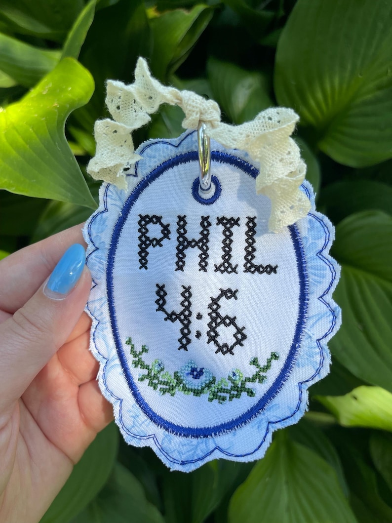 K&ouml;nnte beinhalten: Ein ovales Stoffornament mit dem in schwarzem Kreuzstich gestickten Text PHIL 4:6 &uuml;ber einer kleinen gr&uuml;nen und blauen Blumenstickerei. Der wei&szlig;e Stoff hat einen gewellten blauen Rand und ist mit einem cremefarbenen Spitzenband versehen.