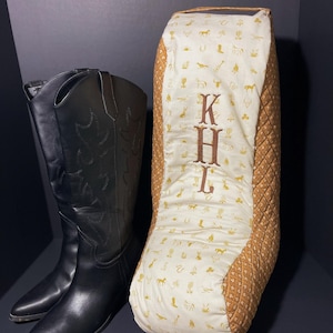 May include: Black leather cowboy boots and a boot bag. The bag is tan and cream, with a quilted pattern and a cream front featuring embroidered letters "KHL" and western-themed icons. The boots are black.