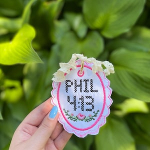 K&ouml;nnte beinhalten: Ein ovales wei&szlig;es Stoffornament mit schwarzem Kreuzstich-Text, der PHIL 4:13 lautet. Das Ornament hat einen rosa gewellten Rand, ein kleines Blumenmuster in Rosa und Gr&uuml;n sowie ein cremefarbenes Spitzenband, das an einem goldenen Ring befestigt ist.