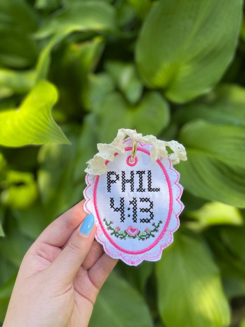 K&ouml;nnte beinhalten: Ein ovales wei&szlig;es Stoffornament mit schwarzem Kreuzstich-Text, der PHIL 4:13 lautet. Das Ornament hat einen rosa gewellten Rand, ein kleines Blumenmuster in Rosa und Gr&uuml;n sowie ein cremefarbenes Spitzenband, das an einem goldenen Ring befestigt ist.