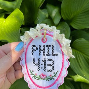 K&ouml;nnte beinhalten: Ein ovales wei&szlig;es Stoffornament mit schwarzem Kreuzstich-Text, der PHIL 4:13 lautet. Das Design umfasst einen rosa gestickten Rand, ein kleines Blumenmotiv am unteren Rand und eine ger&uuml;schte cremefarbene Spitzenborte mit einem goldenen Metallring.