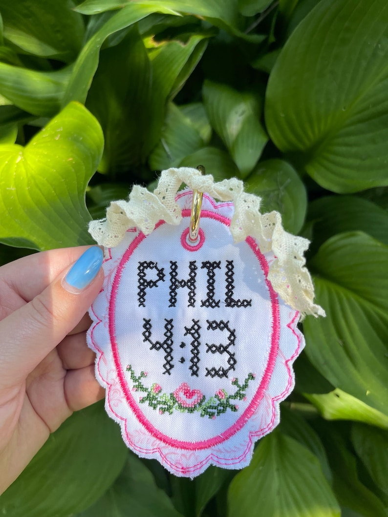 K&ouml;nnte beinhalten: Ein ovales wei&szlig;es Stoffornament mit schwarzem Kreuzstich-Text, der PHIL 4:13 lautet. Das Design umfasst einen rosa gestickten Rand, ein kleines Blumenmotiv am unteren Rand und eine ger&uuml;schte cremefarbene Spitzenborte mit einem goldenen Metallring.