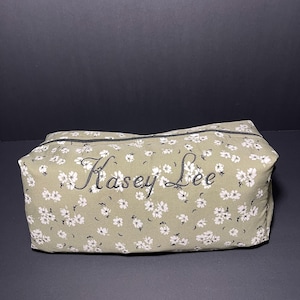 May include: A rectangular cosmetic bag with a floral pattern in white and black on a light green background. The name "Kasey Lee" is embroidered in black script across the top. The bag has a black zipper.