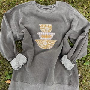 May include: A gray sweatshirt with a crew neck and long sleeves. The cuffs are lined with a soft, gray material. The front of the sweatshirt features a design of three yellow floral and gingham patterned mixing bowls stacked on top of each other.