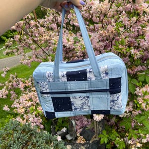 May include: A light blue quilted tote bag with a patchwork design featuring white floral patterns and navy blue accents. The bag has two top handles and a zipper closure.