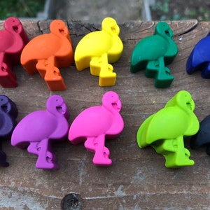 May include: A selection of ten flamingo-shaped wax crayons in a range of colours, including red, orange, yellow, green, blue, purple, pink, light green, and black. The crayons are displayed on a weathered wooden surface.