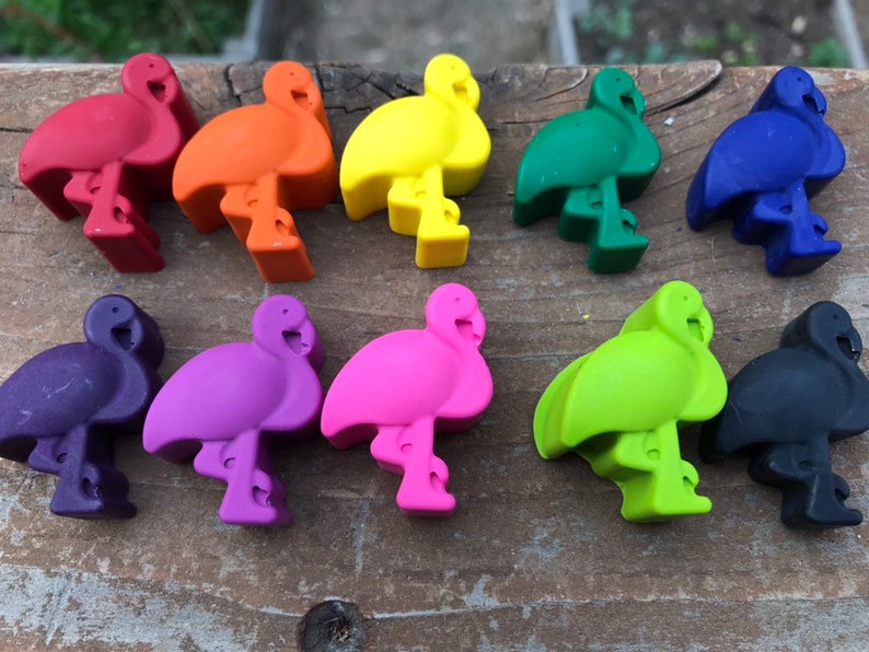 May include: A selection of ten flamingo-shaped wax crayons in a range of colours, including red, orange, yellow, green, blue, purple, pink, light green, and black. The crayons are displayed on a weathered wooden surface.
