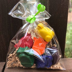 Puede incluir: Una bolsa transparente de celof&aacute;n atada con una cinta verde, llena de ceras de colores con forma de animales. Las ceras son rojas, naranjas, amarillas, verdes, azules y moradas. La bolsa est&aacute; llena de virutas de papel marr&oacute;n.