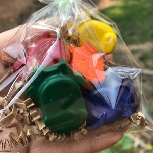 Puede incluir: Una bolsa transparente atada con una cinta verde, llena de ceras de colores con forma de dinosaurio. Las ceras son de color rojo, naranja, amarillo, verde y azul, y descansan sobre papel arrugado marr&oacute;n.