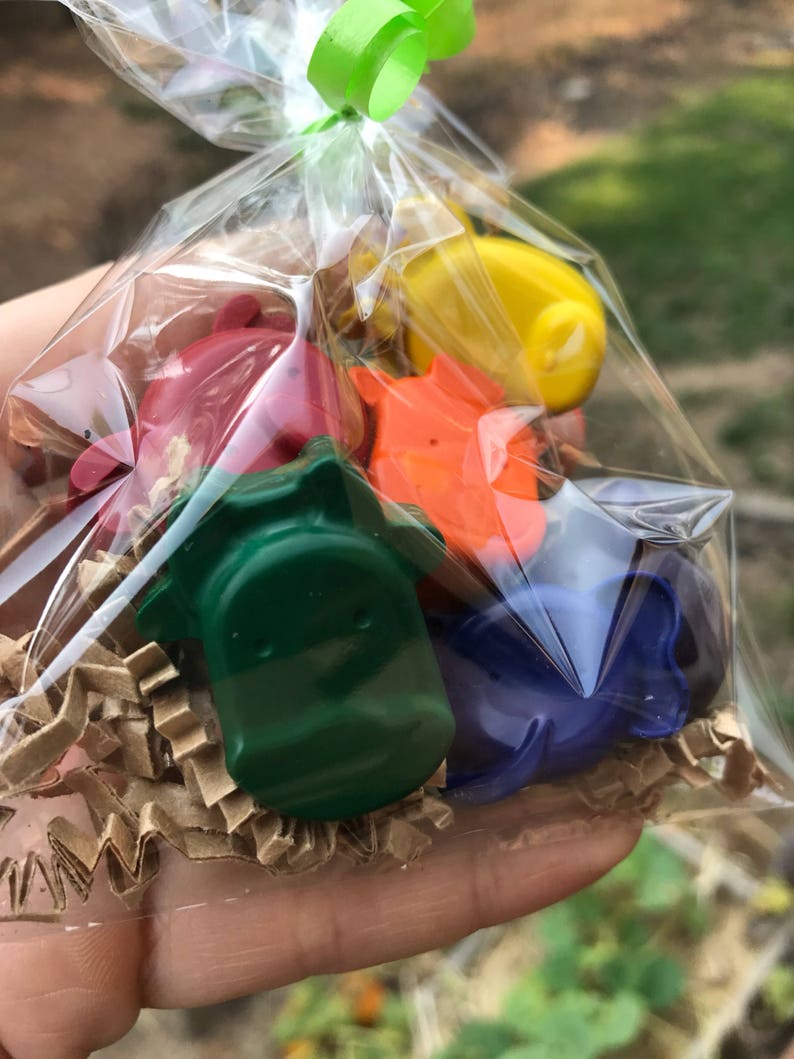Puede incluir: Una bolsa transparente atada con una cinta verde, llena de ceras de colores con forma de dinosaurio. Las ceras son de color rojo, naranja, amarillo, verde y azul, y descansan sobre papel arrugado marr&oacute;n.