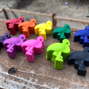 May include: A selection of flamingo-shaped crayons in a range of colours: red, orange, yellow, green, blue, purple, pink, and black. The crayons are displayed on a wooden surface, highlighting their bright hues and distinctive design.
