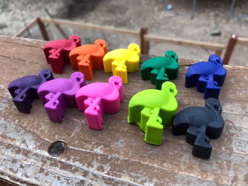 May include: A selection of flamingo-shaped crayons in a range of colours: red, orange, yellow, green, blue, purple, pink, and black. The crayons are displayed on a wooden surface, highlighting their bright hues and distinctive design.